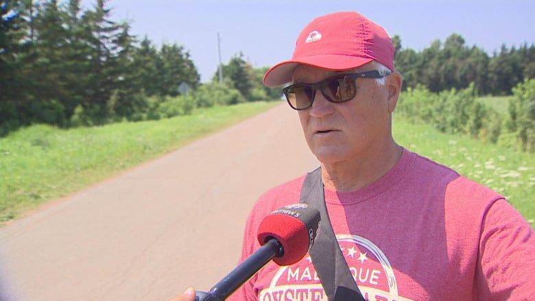 A man in a red shirt stands on a dirt road, speaking into a microphone.