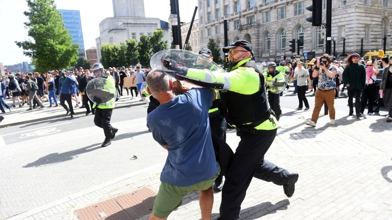 A police officer with a baton and shield clashes with a person as a crowd of people look on.