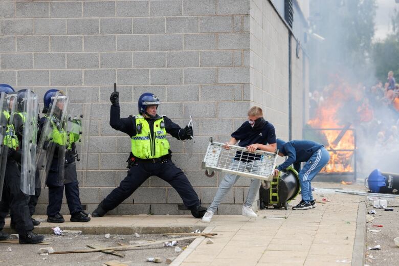 A police officer in front of a line of other officers swings a baton at protesters holding objects as a fire burns in the background.