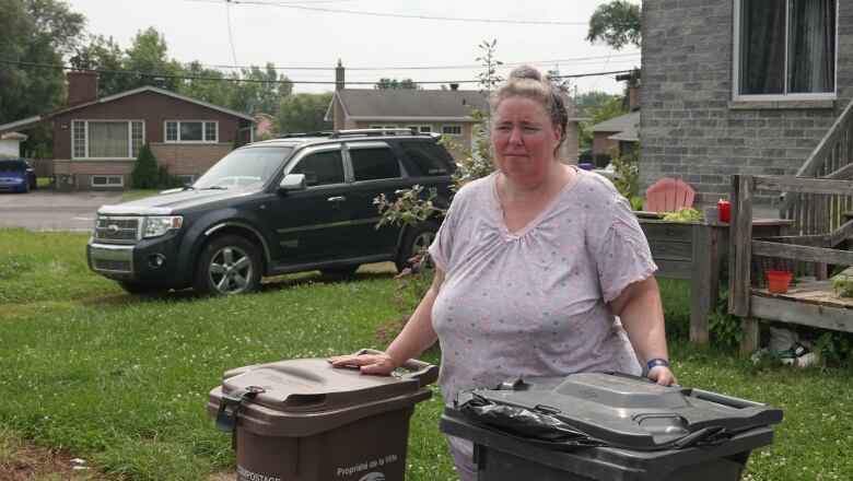 A blonde woman stands with her hands on two garbage cans.