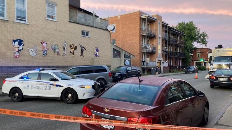 Police cars and tape block off a section of a street with apartments. 