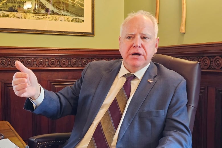 A man in a suit gestures and talks as he sits in a chair.