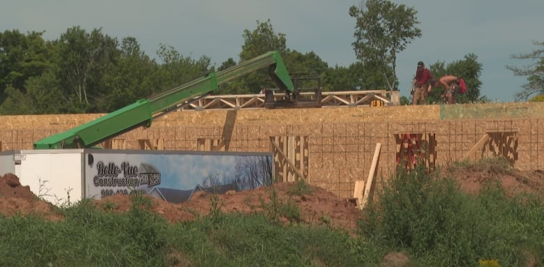 Two men are seen working on the roof of a new wooden building, with a construction crane in the foreground. 