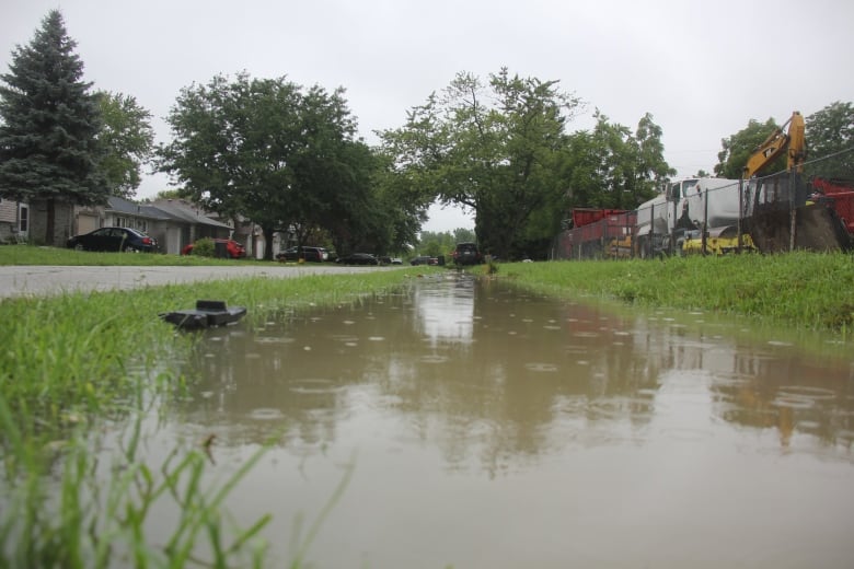 A ditch filled with muddy brown water on the side of a roadway. Droplets are visible hitting the surface of the water in the ditch due to continued rain.