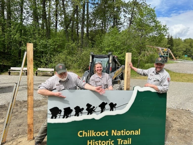 Three people standing behind a sign that says 'Chilkoot National Historic Trail'