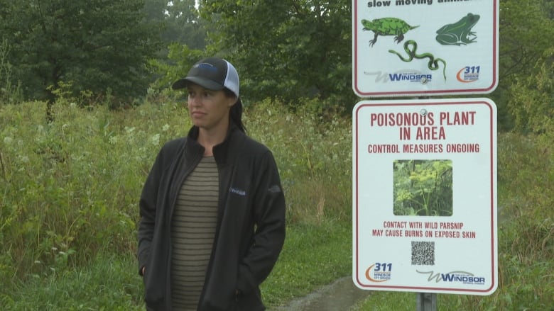 A woman in a baseball cap and rain jacket stand next to a sign warning of invasive yellow parsnips. Behind her is a fielded area