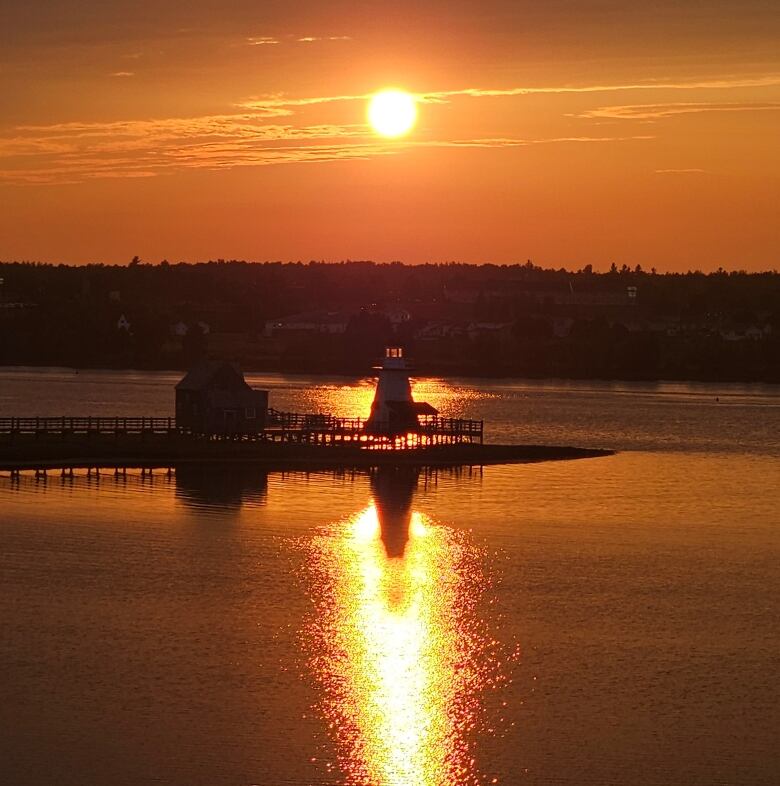 Light reflects over a bay from the setting sun, a lighthouse directly in its path.