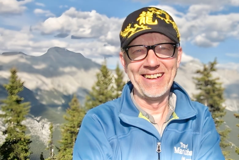 A man in a black cap and blue sweater stands before a mountain. 