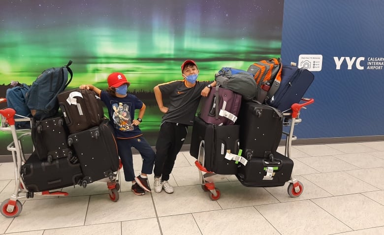 Two boys wearing face masks stand by luggage trolleys stacked with suitcases. 