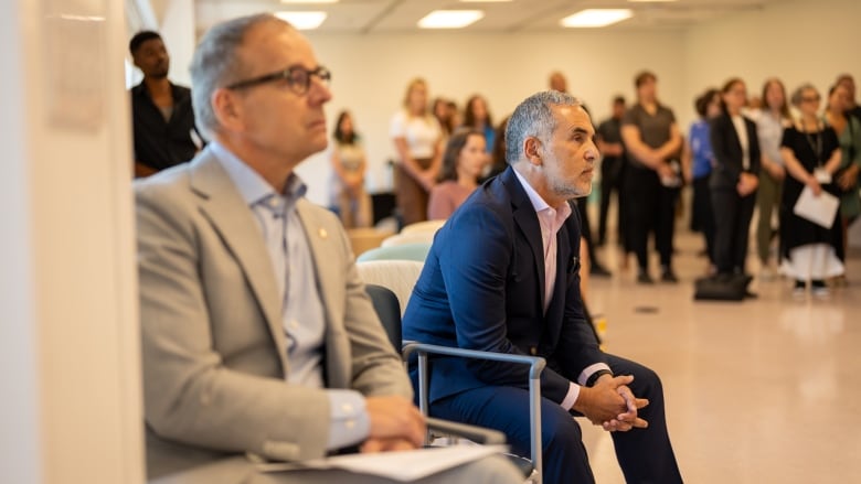 Two men in suits sit in a meeting room on chairs. The crowd behind them is standing. Everyone's attention is directed somewhere out of frame.