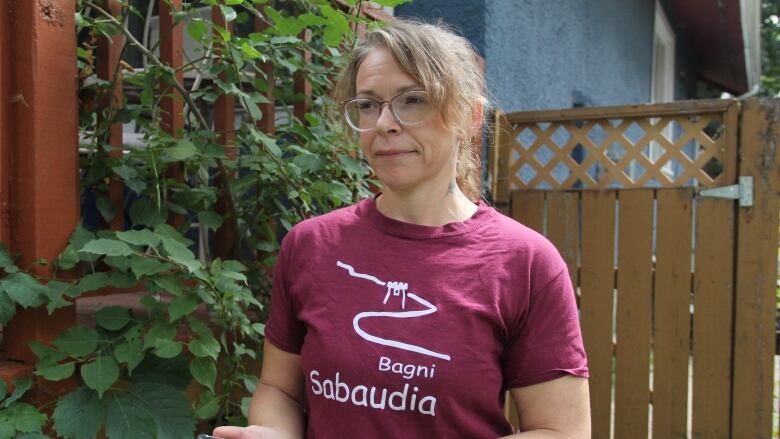 A woman in a burgundy t-shirt looks forward while holding her cell-phone. She's standing in front of a fence with green plants hanging nearby.