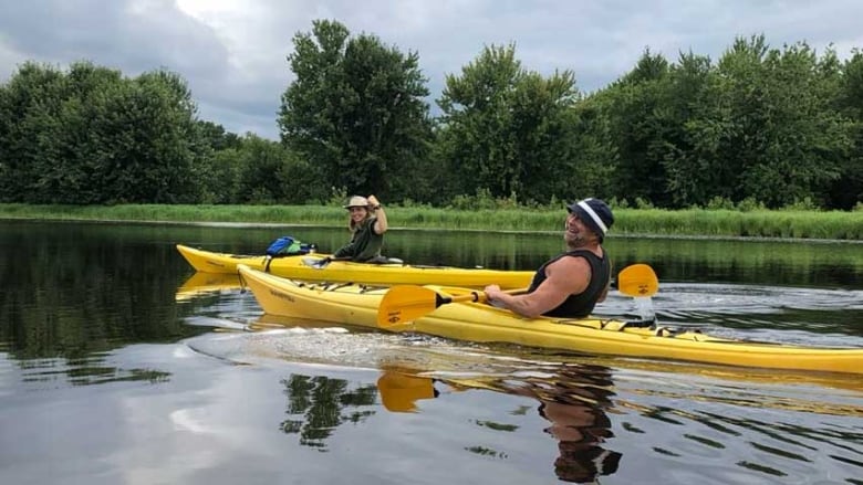 Two people in yellow sea kayaks on calm water with trees in the background.