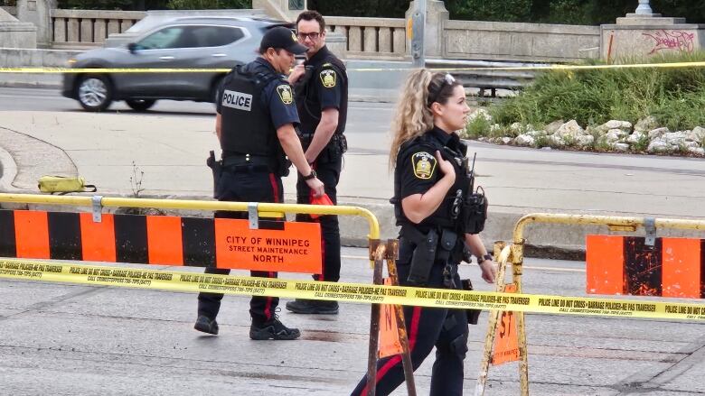 Police officers in black uniforms walk among yellow tape and barricades on a street.