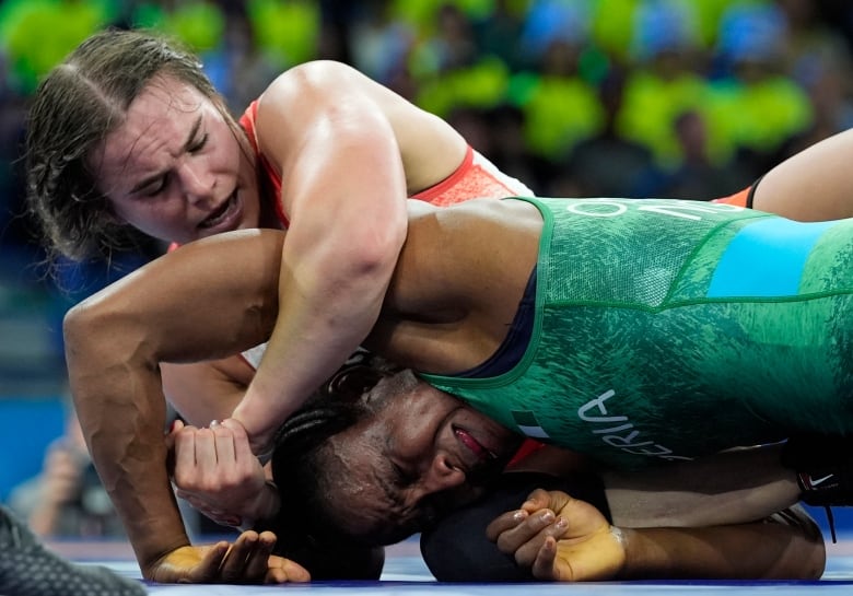 A woman with brown hair wrestles another woman in a green wrestling singlet. The woman with brown hair is on top of the other woman, and her arm is wrapped around the other's arm in a hold.