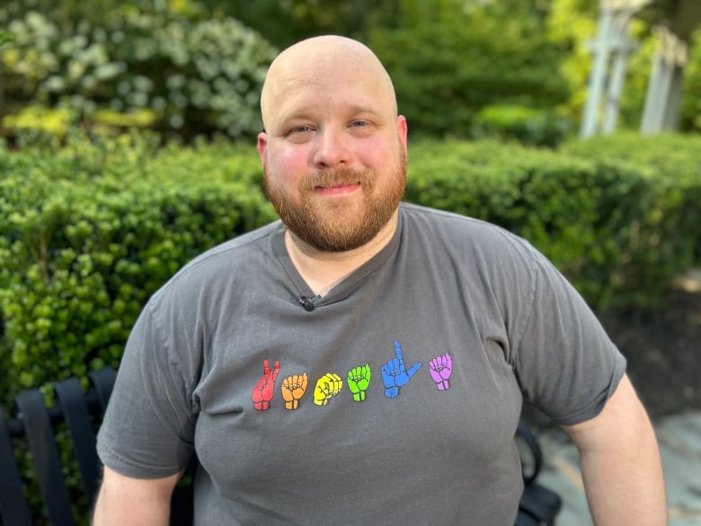 Piece Hacking is a larger man with a bald head and auburn beard and moustache. He is smiling. He has on a gray shirt with sign language on the front in the colors of the rainbow.