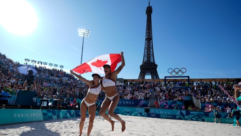 Two beach volleyball athletes celebrate with the Canadian flag in the sand as the Eiffel Tower and the Olympic logo is seen behind them.