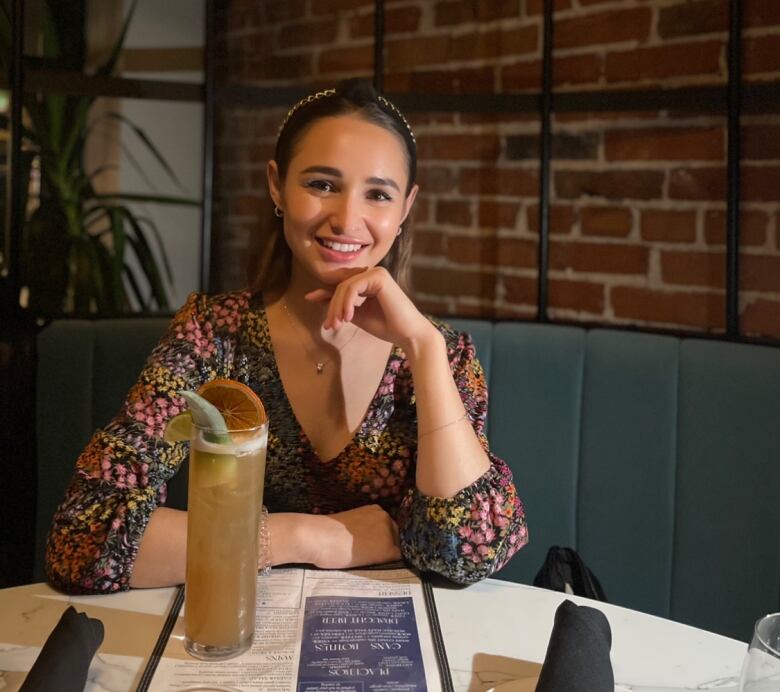 A woman smiles, in front of a brick backdrop with a drink in front of her.