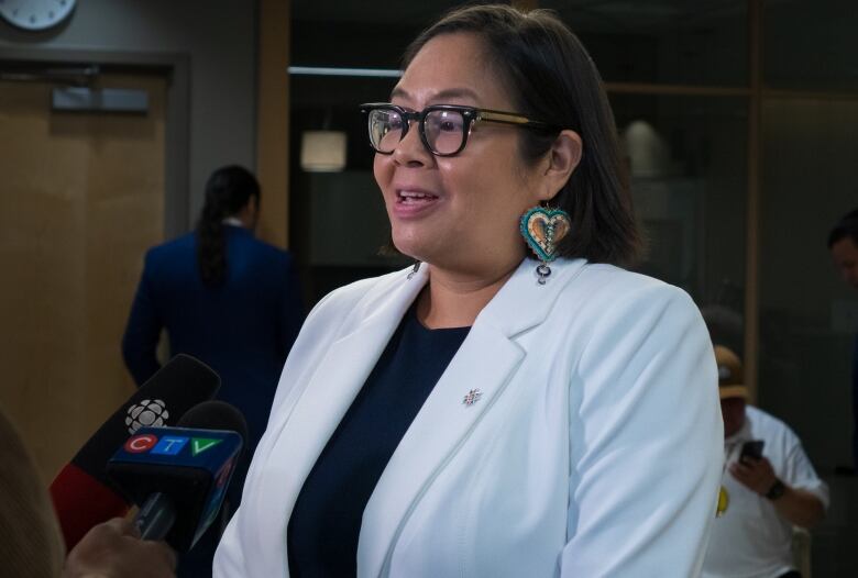 An Indigenous woman wearing a white blazer and beaded earrings shaped like hearts speaks into two media microphones.