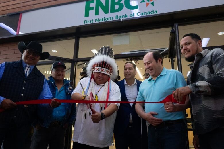 An Indigenous man wearing a headdress uses a pair of oversized scissors to cut a ribbon in front of a set of glass doors, with other people around him