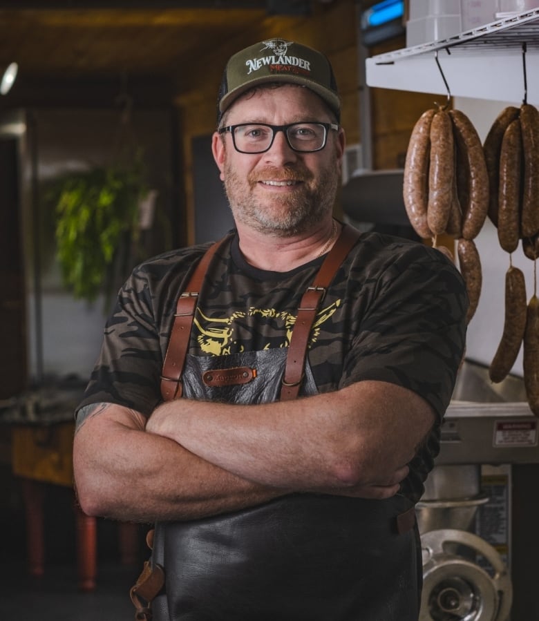 Man with arms folded in a kitchen, a string of sausages hangs behind him.