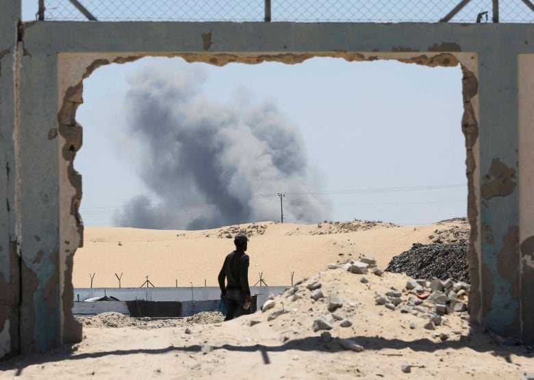 Grey smoke rises over the sandy horizon. In the foreground, a silhouetted figure is framed by what appears the gaping hole is a damaged wall.