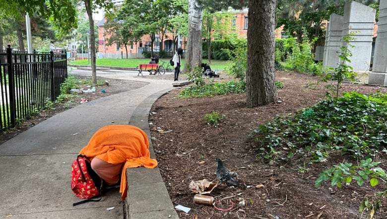 A person, covered with an orange blanket, sits slumped over on a ledge in a park. Litter is seen on the ground nearby.