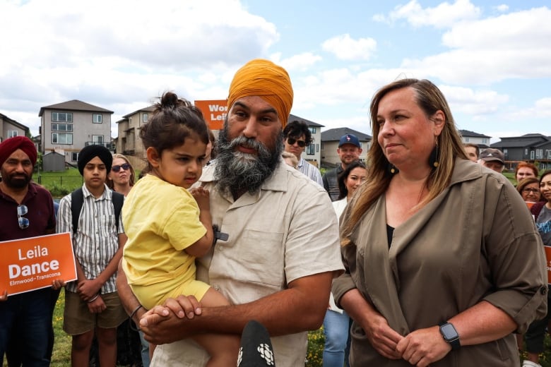 A man holding a baby stands next to a woman, in front of a group of people.