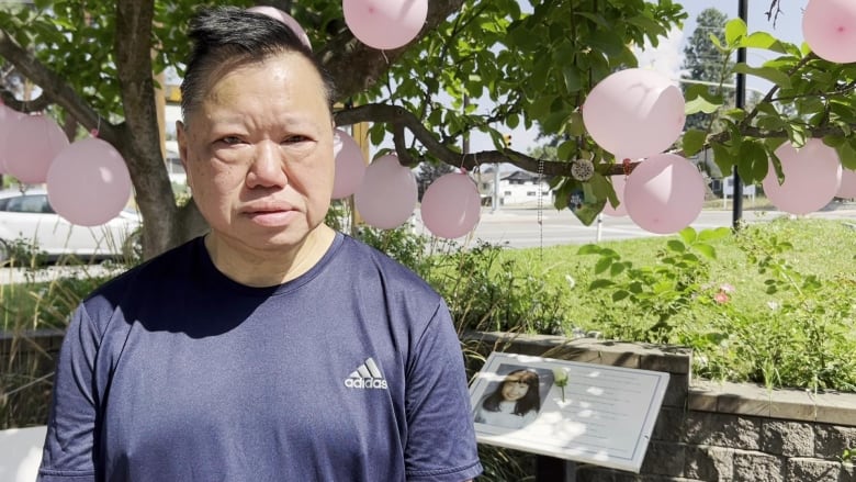 An East Asian man looks forlorn as he stands outdoors in front of a plaque and a series of pink balloons.