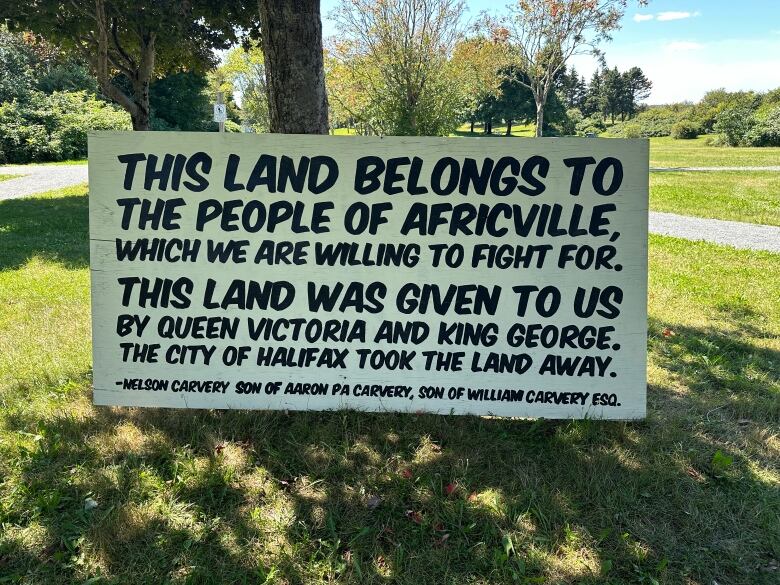 A white sign with black writing leans against a tree. It reads