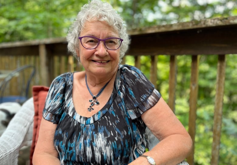 A woman in a blue T-shirt and glasses poses for a picture on a deck with leafy green trees in the background.