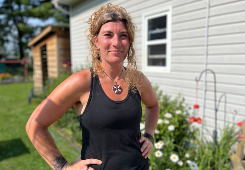 A woman with curly hair in a bun wearing a black tank top poses for a photo outside a duplex with a garden in front.