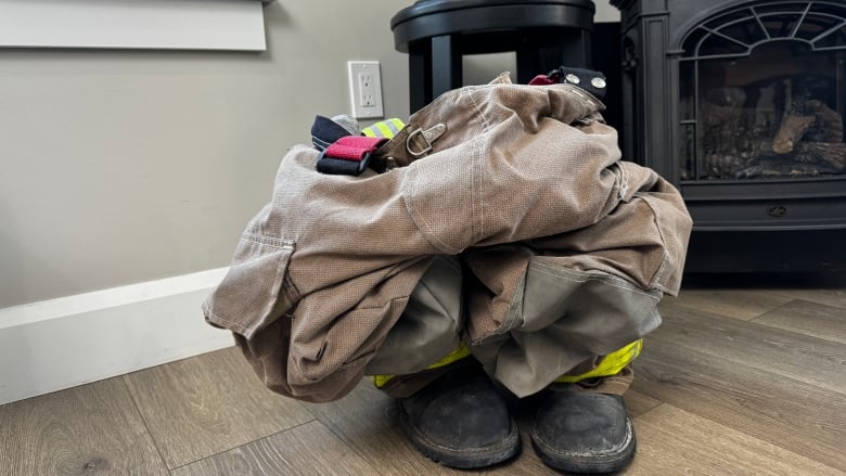 Brown coverall pants are shown crumpled to the floor on top of black boots near a woodstove. A black memorial fire hat is shown in the foreground.