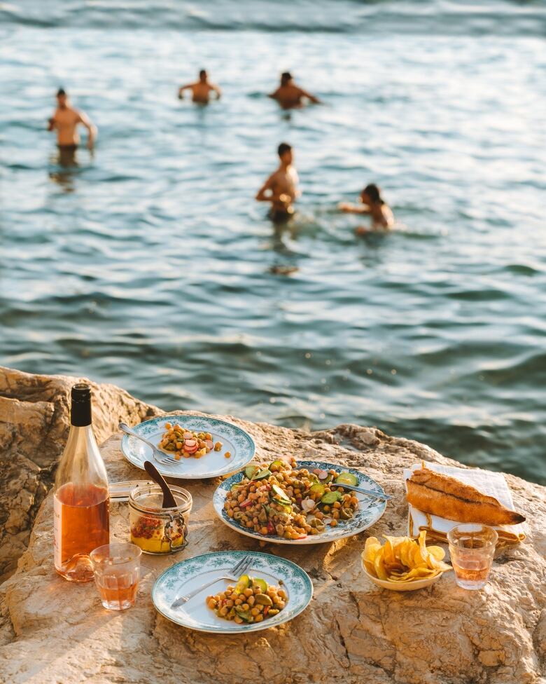 Closeup on a picnic spread of plates of chickpea salad, wine, baguette and a bowl of chips on a large rock at the beach. People swim in the sea in the background. 