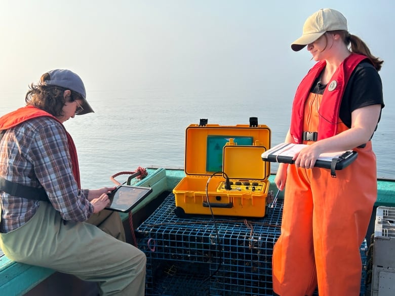 Two women on a fishing boat. One working on a table while other holding a notepad.