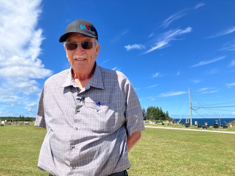 An elderly man in a blue shirt with a black hat and sunglasses in front of a cemetery. 