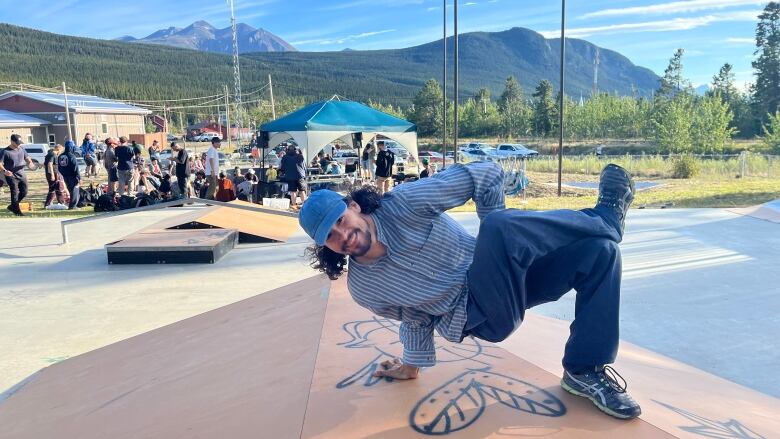 A man with long hair and blue baseball cap folds his body into a pretzel shape on top of a skateboard ramp.