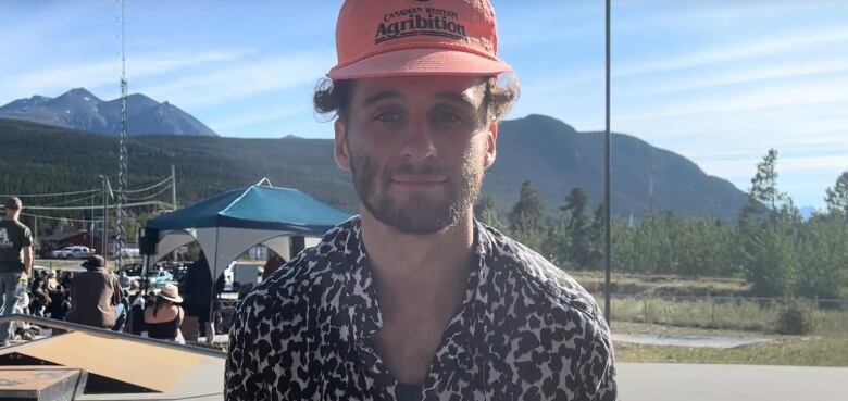 A man with a black white leopard printed button up shirt, orange baseball cap and a beard sits on a skateboard ramp