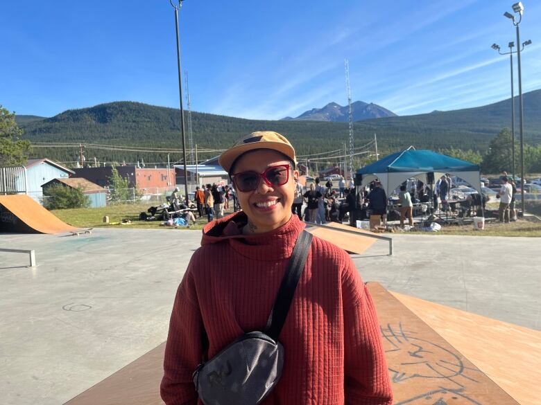 A lady is a red sweater, crossbody bag, yellow hat and sunglasses stands next to a skatepark ramp.