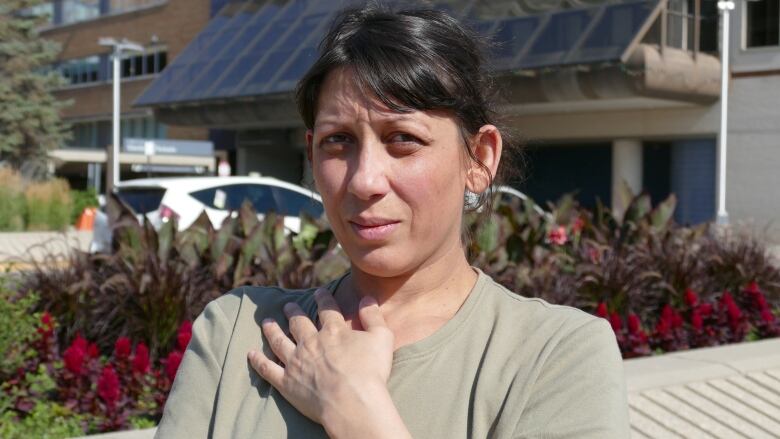 A woman stands outside posing with her left hand laying on her chest, near her right shoulder. 