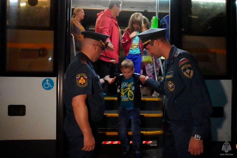 A young boy walks down the stairs of what appears to be a bus, helped down to the ground level by two men with police-like uniforms.