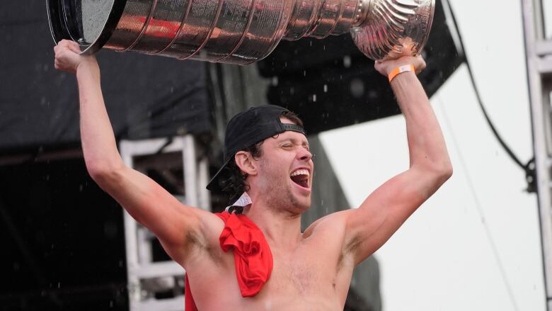 A topless person holds a big silver trophy above their head.