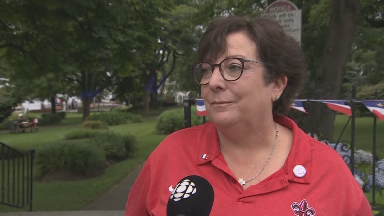 woman in red fleur de lis shirt stands near a railing lined with Acadian flags.