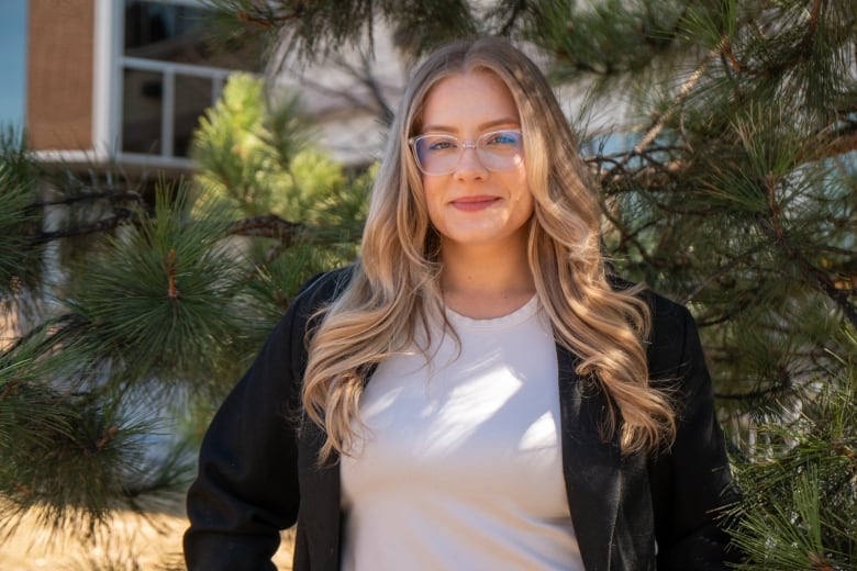 A person with long blonde hair and glasses is seen standing in front of a tree.