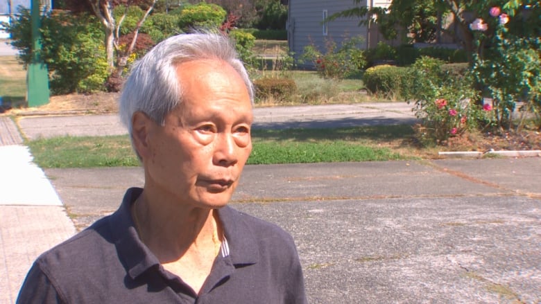 A man with white hair and an open-collared shirt stands on a sidewalk, speaking to someone off-camera.