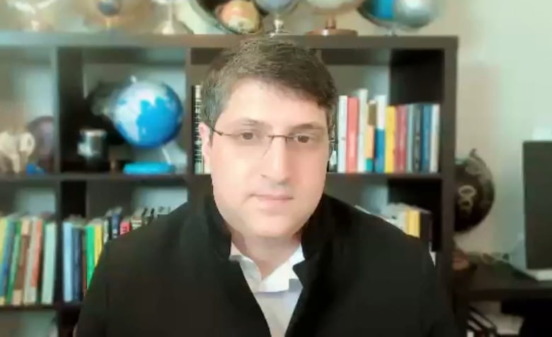 A man in a black jacket and white shirt is on a video call in front of a bookshelf with globes.