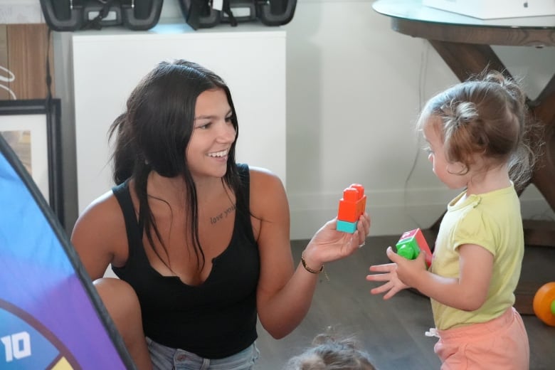 A woman sits on the ground, holding up colourful blocks to hand to a little girl standing in front of her. The girl has her hair in side ponytails and is looking at the woman.