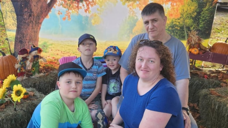 A woman in a blue dress, next to a boy in blue and green t-shirt stand in front of a boy in a blue hat and blue t shirt, and a boy in a black t shirt, and a man in a grey shirt. 