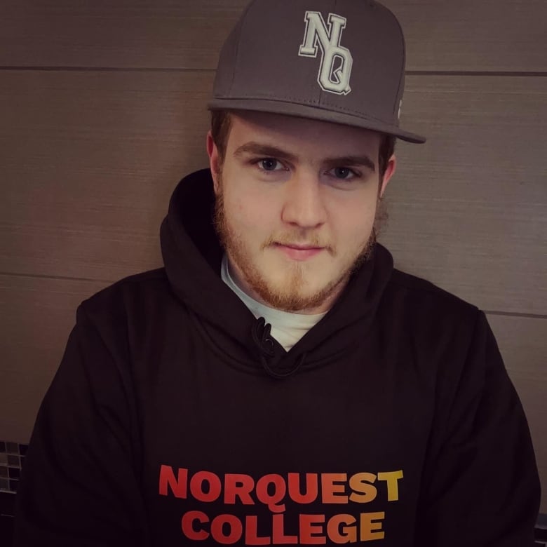 A young man with a light brown beard wears a black baseball cap with the letters NQ on it, and a black sweatshirt that says 