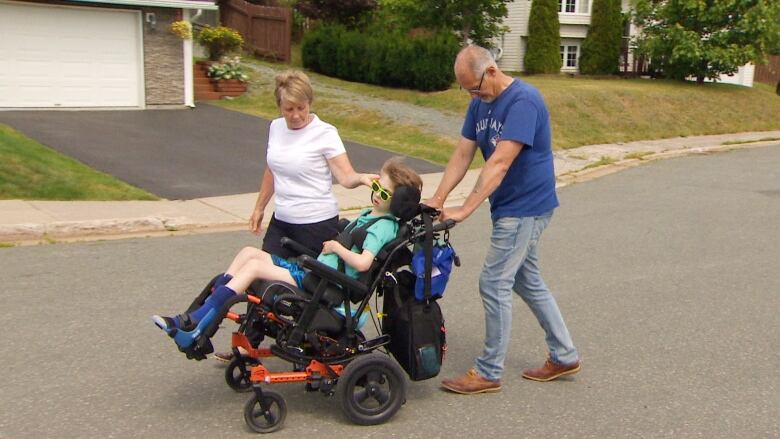 Older woman and man push wheelchair with young child in it on a residential street.