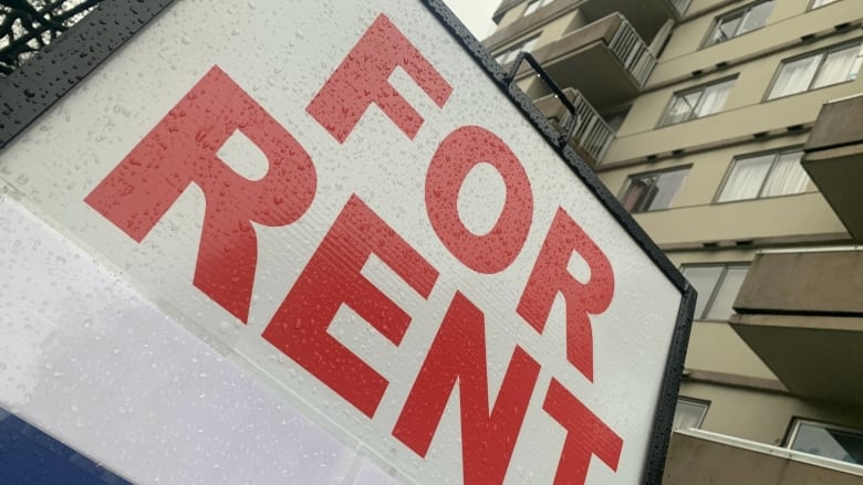 A sign with red letters reads FOR RENT. It's wet with rain, on an angle, and there's a beige highrise apartment building filling out the background.